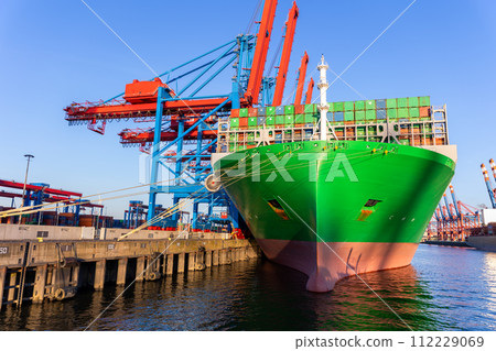 Scenic view giant cargo container ship loading Hamburg city port harbor against seaport cranes warm evening sunset lights. Global commercial trade freight charter shipping and logistics background Scenic view giant cargo container ship loading Hamburg city port harbor against seaport cranes warm evening sunset lights. Global commercial trade freight charter shipping and logistics background 112229069