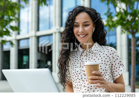 A young professional woman takes a relaxing coffee break outdoors, using her laptop against the backdrop of a modern office building. This candid moment captures the balance between work A young professional woman takes a relaxing coffee break outdoors, using her laptop against the backdrop of a modern office building. This candid moment captures the balance between work 112229085