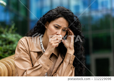 A distressed businesswoman in professional attire is sitting outside, wiping away tears with a tissue, a look of sadness and worry on her face amidst an urban backdrop. 112229096
