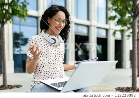 A young female professional engages in a video call while working on her laptop outside a modern office building, portraying flexibility and modern work lifestyle. A young female professional engages in a video call while working on her laptop outside a modern office building, portraying flexibility and modern work lifestyle. 112229098