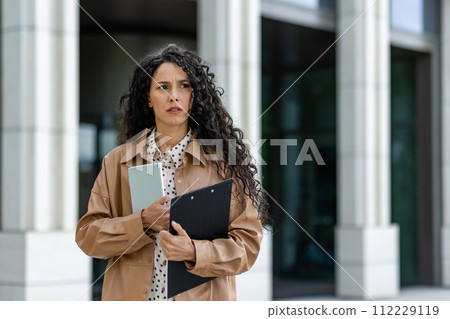 A professional woman stands outside a modern building, poised with a tablet and clipboard in hand. Her fashionable trench coat and smart attire suggest business and efficiency in an urban setting. 112229119