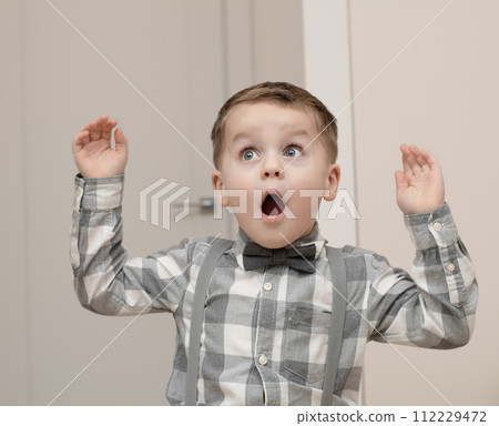 Emotions of surprise and delight on the child s face. A small handsome boy of 4 years old in a shirt with a bow tie shows a grimace with an expression on his face. Close-up. Emotions of surprise and delight on the child s face. A small handsome boy of 4 years old in a shirt with a bow tie shows a grimace with an expression on his face. Close-up. 112229472