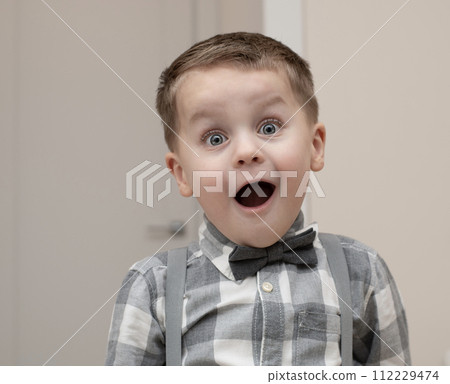Emotions of surprise and delight on the child s face. A small handsome boy of 4 years old in a shirt with a bow tie shows a grimace with an expression on his face. Close-up. Emotions of surprise and delight on the child s face. A small handsome boy of 4 years old in a shirt with a bow tie shows a grimace with an expression on his face. Close-up. 112229474