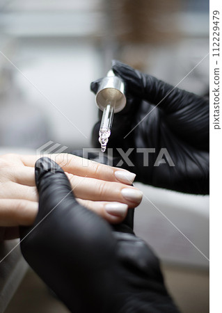 Beauty concept. A manicurist in black latex gloves makes a hygienic manicure and paints the client's nails with gel polish and drips oil on the cuticles in a beauty salon. 112229479