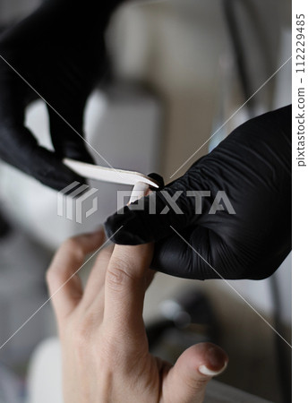 Beauty concept. A manicurist in black latex gloves makes a hygienic manicure, paints the client's nails with gel polish and files them with a nail file in a beauty salon. 112229485