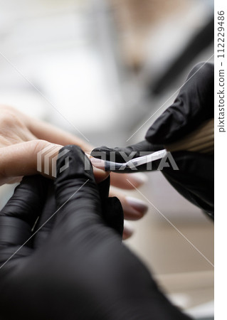 Beauty concept. A manicurist in black latex gloves makes a hygienic manicure and paints the client's nails with gel polish in a beauty salon. Close-up. Beauty concept. A manicurist in black latex gloves makes a hygienic manicure and paints the client's nails with gel polish in a beauty salon. Close-up. 112229486