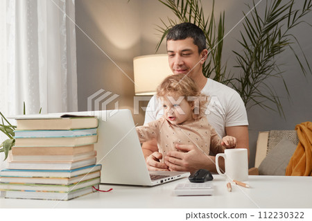 Smiling cheerful Caucasian man wearing casual clothing sitting at table in front of laptop with his infant baby daughter watching cartoons online together in home interior 112230322