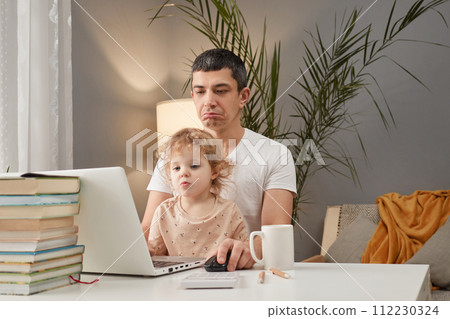 Disappointed sad upset Caucasian man wearing casual clothing sitting at table in front laptop with his little daughter of in home interior being unhappy to loose level of video game 112230324