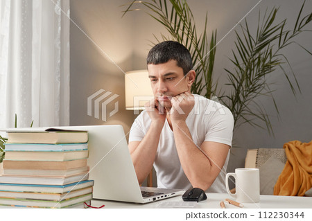 Bored sad Caucasian adult man wearing white T-shirt working at home with laptop looking at computer display with negative feeling has no mood and desire to work Bored sad Caucasian adult man wearing white T-shirt working at home with laptop looking at computer display with negative feeling has no mood and desire to work 112230344