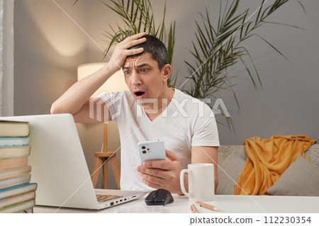 Shocked scared Caucasian man working on laptop and looking at display making facepalm gesture and holding mobile in hands sitting at workplace in home interior 112230354