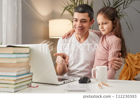 Smiling Caucasian man wearing casual clothing sitting at table in front of computer in home interior with his brown haired daughter enjoying digital time spending together Smiling Caucasian man wearing casual clothing sitting at table in front of computer in home interior with his brown haired daughter enjoying digital time spending together 112230383