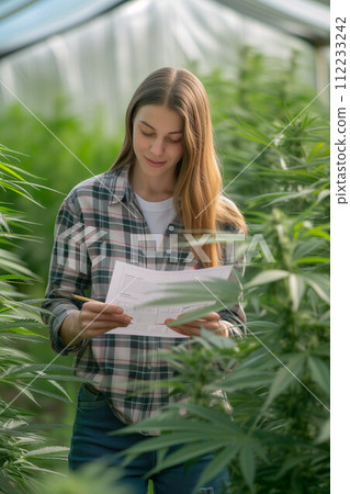 Young Caucasian woman holds documents to examine marijuana plants in outdoor farm. Young Caucasian woman holds documents to examine marijuana plants in outdoor farm. 112233242