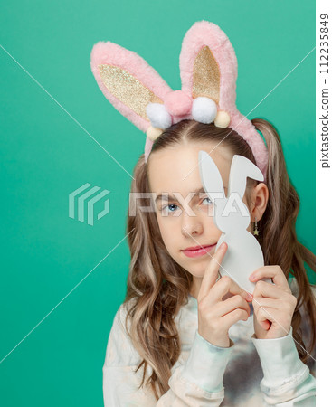 Girl with happy smile, wearing shiny pink bunny ears holding wooden bunny figurine, blue background, Easter fun. 112235849