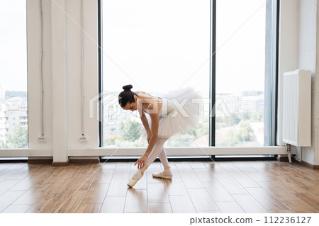 Ballerina stretching to feet while standing in dance studio with large windows. Ballerina stretching to feet while standing in dance studio with large windows. 112236127