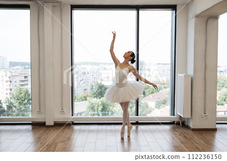 Ballerina practice ballet positions in tutu skirt near window in white hall. 112236150