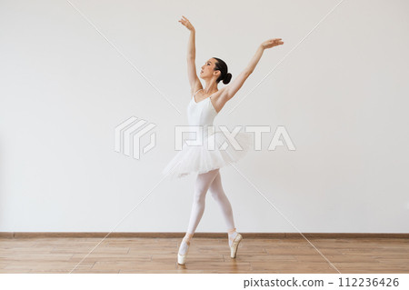 Caucasian woman ballerina in white tutu, dancing on pointe with arms overhead. 112236426