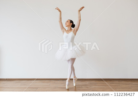 Caucasian woman ballerina in white tutu, dancing on pointe with arms overhead. 112236427