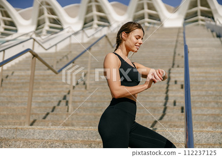Smiling woman in sportswear looking on smartwatch after training. Outdoor sports in the morning 112237125