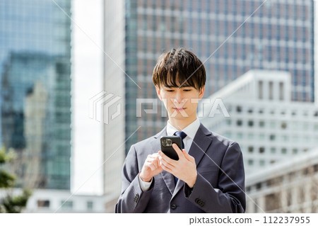 一名 20 多歲的西裝男子在大城市市中心手持智慧型手機實施網路犯罪。 112237955