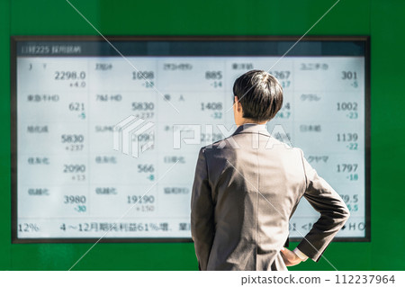 Rear view of a businessman in his 30s in a suit, with his hands on his hips, looking at the stocks on the electronic bulletin board 112237964