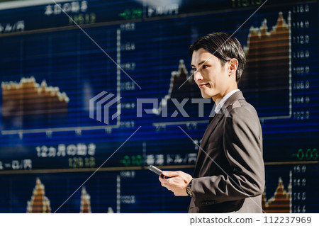 A man in his 30s predicts stock prices in front of an electronic bulletin board and makes money using his smartphone. 112237969