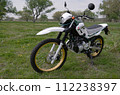 A parked off-road bike receives light from the opposite side from the bright clouds through which the setting sun shines through in a meadow on a spring evening. 112238397