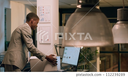 Sad african american employee packing desk belongings after being laid off from job after redundant staff reduction. Businessman putting office decorations in box after being fired Sad african american employee packing desk belongings after being laid off from job after redundant staff reduction. Businessman putting office decorations in box after being fired 112239289
