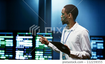 African american specialist using tablet to check data center security features protecting against unauthorized access, data breaches, DDoS attacks and other cybersecurity threats 112239355