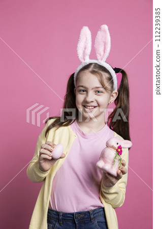 Optimistic small kid posing with painted eggs and a pink rabbit toy, wearing fluffy bunny ears and being cheerful in front of camera. Young girl with pigtails showing her easter decorations. Optimistic small kid posing with painted eggs and a pink rabbit toy, wearing fluffy bunny ears and being cheerful in front of camera. Young girl with pigtails showing her easter decorations. 112239385