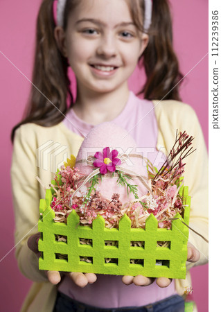 Positive excited small kid showing her handmade easter eggs in a colored basket, feeling proud of her arrangement. Young child with bunny ears holding festive decorations, april celebrations. Positive excited small kid showing her handmade easter eggs in a colored basket, feeling proud of her arrangement. Young child with bunny ears holding festive decorations, april celebrations. 112239386