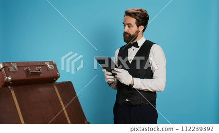 Classy doorman porter reviewing tablet, in charge of baggage for safekeeping. Bellboy presenting his commitment in providing outstanding concierge service, symbolizing his occupation at chic hotel. 112239392