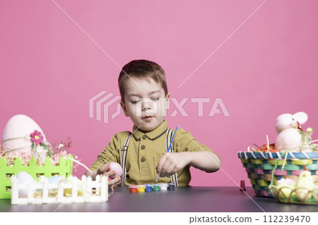 Delighted young kid coloring eggs and ornaments with watercolors as he prepares artistic creations for Easter Sunday festivities. Playful toddler having fun in the studio with tie dye painting. Delighted young kid coloring eggs and ornaments with watercolors as he prepares artistic creations for Easter Sunday festivities. Playful toddler having fun in the studio with tie dye painting. 112239470