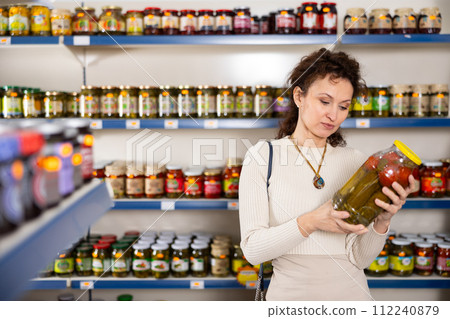 In Russian goods store, woman choose jar of pickled tomatoes and cucumbers 112240879