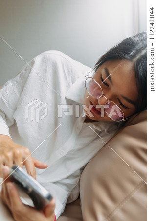 Side view of asian Thai office woman using mobile phone while leaning and resting on brown couch sofa, tired of work. Side view of asian Thai office woman using mobile phone while leaning and resting on brown couch sofa, tired of work. 112241044