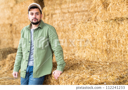 Male farmer posing against the background of hay bales in hayloft 112241233