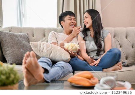 Happy young Asian couple enjoy eating popcorn and watching a movie while relaxing on a sofa. Happy young Asian couple enjoy eating popcorn and watching a movie while relaxing on a sofa. 112249656