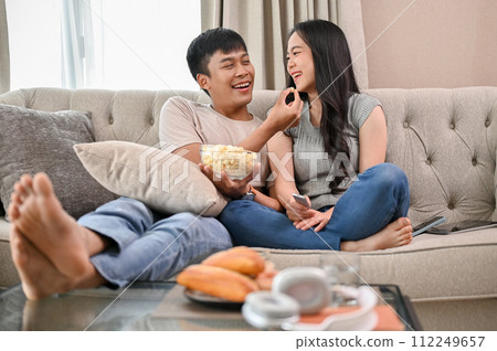 Happy young Asian couple enjoy eating popcorn and watching a movie while relaxing on a sofa. 112249657
