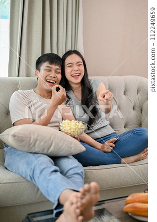 Cheerful Asian couple are enjoying watching a movie, and eating popcorn on a sofa at home together. 112249659