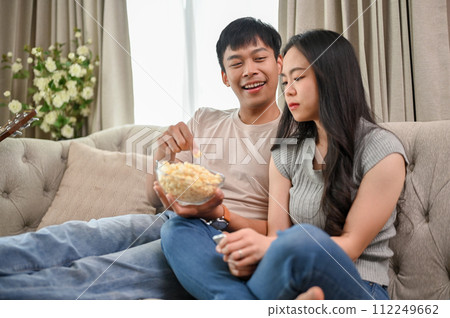 Happy young Asian couple enjoy eating popcorn and watching a movie while relaxing on a sofa. Happy young Asian couple enjoy eating popcorn and watching a movie while relaxing on a sofa. 112249662