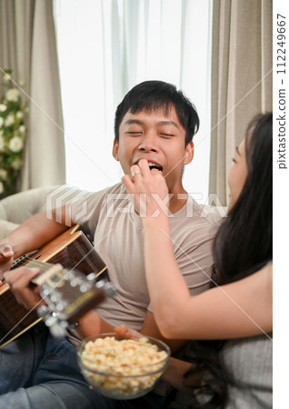 A caring young girlfriend is feeding popcorn to her boyfriend while he plays the guitar. A caring young girlfriend is feeding popcorn to her boyfriend while he plays the guitar. 112249667