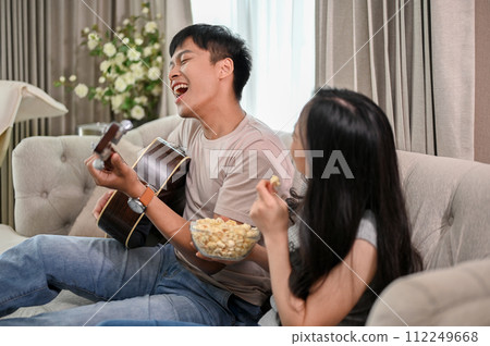 Cheerful boyfriend is playing an acoustic guitar and singing, while his girlfriend dances on a sofa. Cheerful boyfriend is playing an acoustic guitar and singing, while his girlfriend dances on a sofa. 112249668