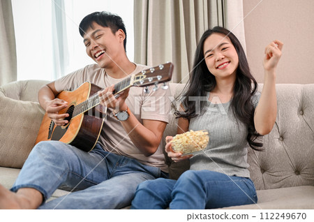 Cheerful boyfriend is playing an acoustic guitar and singing, while his girlfriend dances on a sofa. Cheerful boyfriend is playing an acoustic guitar and singing, while his girlfriend dances on a sofa. 112249670