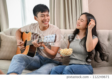 Cheerful boyfriend is playing an acoustic guitar and singing, while his girlfriend dances on a sofa. 112249671