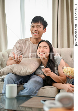 A happy young Asian couple is relaxing on a sofa, enjoying watching television together at home. 112249691