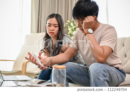 An unhappy young Asian couple is arguing about high domestic bills on a sofa in the living room. An unhappy young Asian couple is arguing about high domestic bills on a sofa in the living room. 112249704