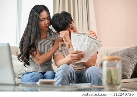 A stressed young married couple is sitting on a sofa, concerned about their invoices. 112249720