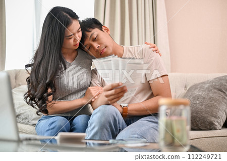 A stressed young married couple is sitting on a sofa, concerned about their invoices. 112249721