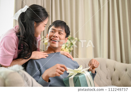 A lovely Asian woman is surprising her husband with a present in the living room. happy anniversary A lovely Asian woman is surprising her husband with a present in the living room. happy anniversary 112249737