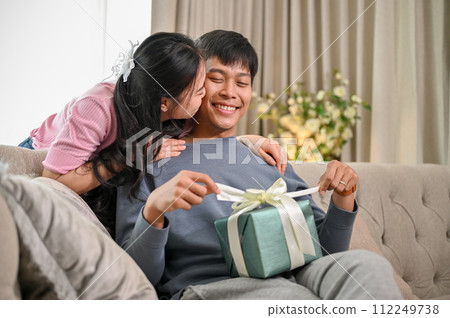 A lovely Asian wife is kissing her husband's cheek on a couch after surprising him with a present. A lovely Asian wife is kissing her husband's cheek on a couch after surprising him with a present. 112249738