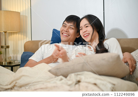 A happy young Asian couple in pajamas is enjoying watching television or a movie together in bed. 112249896